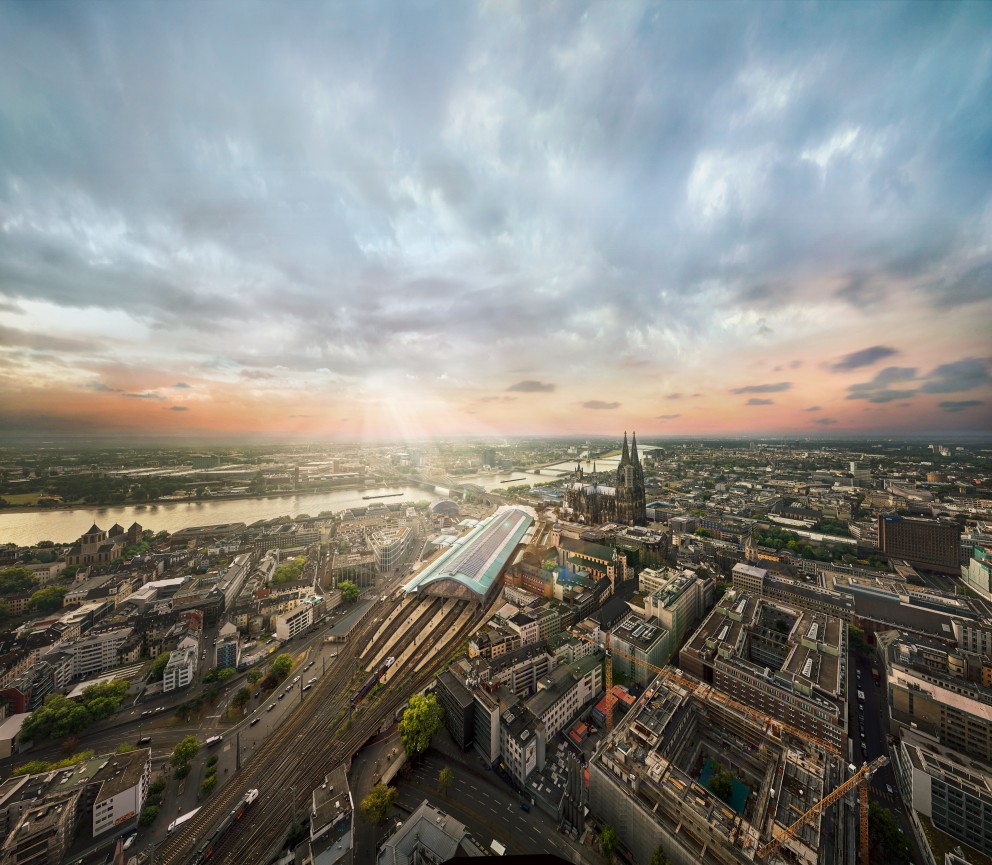 Das Bild zeigt die Stadt Köln von oben. Man sieht im Vordergrund den Kölner Hauptbahnhof. Direkt daneben steht der Kölner Dom. Im Hintergrund fliest der Rhein durch die Stadt. Die Sonne geht auf und erzeugt eine rötliche Himmelsstimmung.