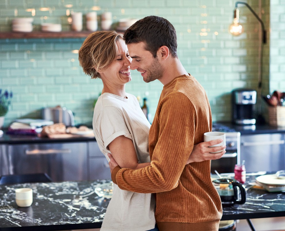 Das Bild zeigt ein junges Pärchen frühmorgens in der Küche. Hinter ihnen steht ein Küchentisch mit einem frühstück. Das junge Paar liegt sich in den Armen und lächelt sich an. Sie hält in Ihrer Hand eine Tasse.