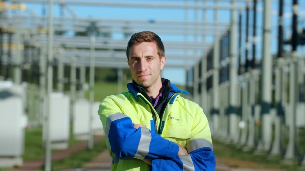 Das Bild zeigt eine Mitarbeiter der Firma Amprion. Er trägt seine Arbeitsschutzkleidung und steht in einer Umspannanlage. Im Hintergrund sieht man mehrere Sammelschienen, worüber der Strom in der Umspannanlage auf andere Leitungen geleitet wird.