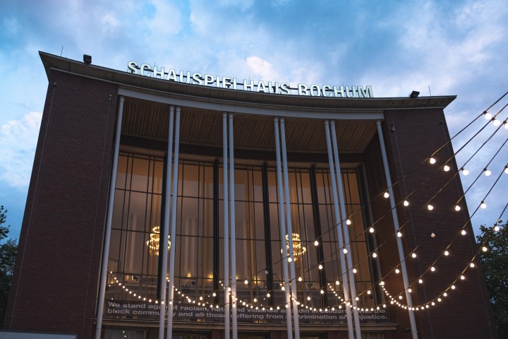 Das Bild zeigt das Schauspielhaus Bochum von Vorne. Während im Hintergrund die Dämmerung eintritt fangen im Vordergrund Girlanden an zu leuchten. Das Geäude ist aus Backsteinen und hat eine Front mit mehreren Fenstern.