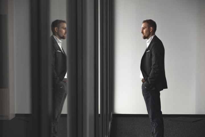 Das Bild zeigt Yannick Jonat vor einer Glaswand. in der Glaswand spiegelt er sich. Yannick Jonat schaut nachdenklich und trägt einen schwarzen Anzug.